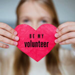 Woman holding a heart-shaped sign reading "Be My Volunteer"