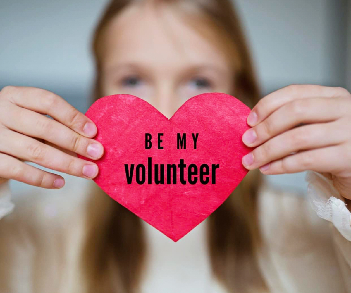 Woman holding a heart-shaped sign reading "Be My Volunteer"
